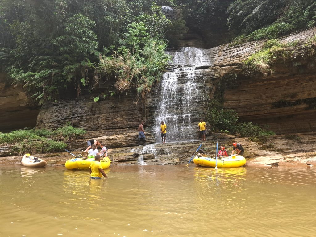 Enjoying a break beneath a cascading waterfall during their Upper Navua River Rafting trip in Fiji, surrounded by lush greenery and rocky cliffs.